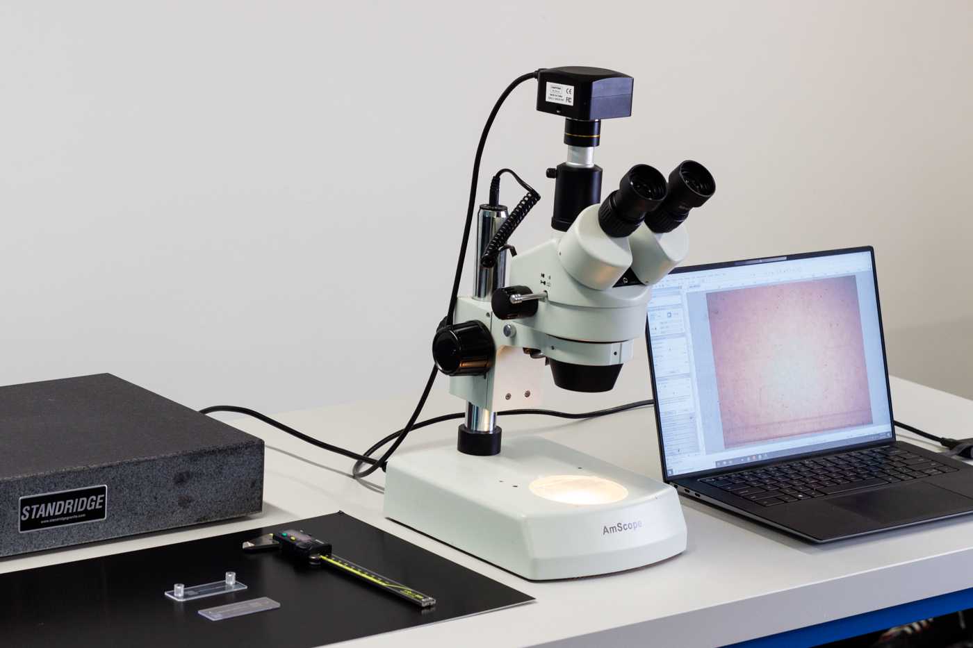 A microscope on a bench top with a laptop to the right and a set of calipers and two microfluidic devices to the left.